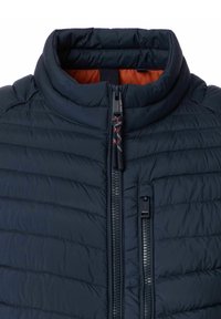 Marineblauwe gewatteerde jas met hoge kraag, voorzien van een dubbele ritssluiting en oranje binnenvoering. Textuur met horizontale stiknaden.