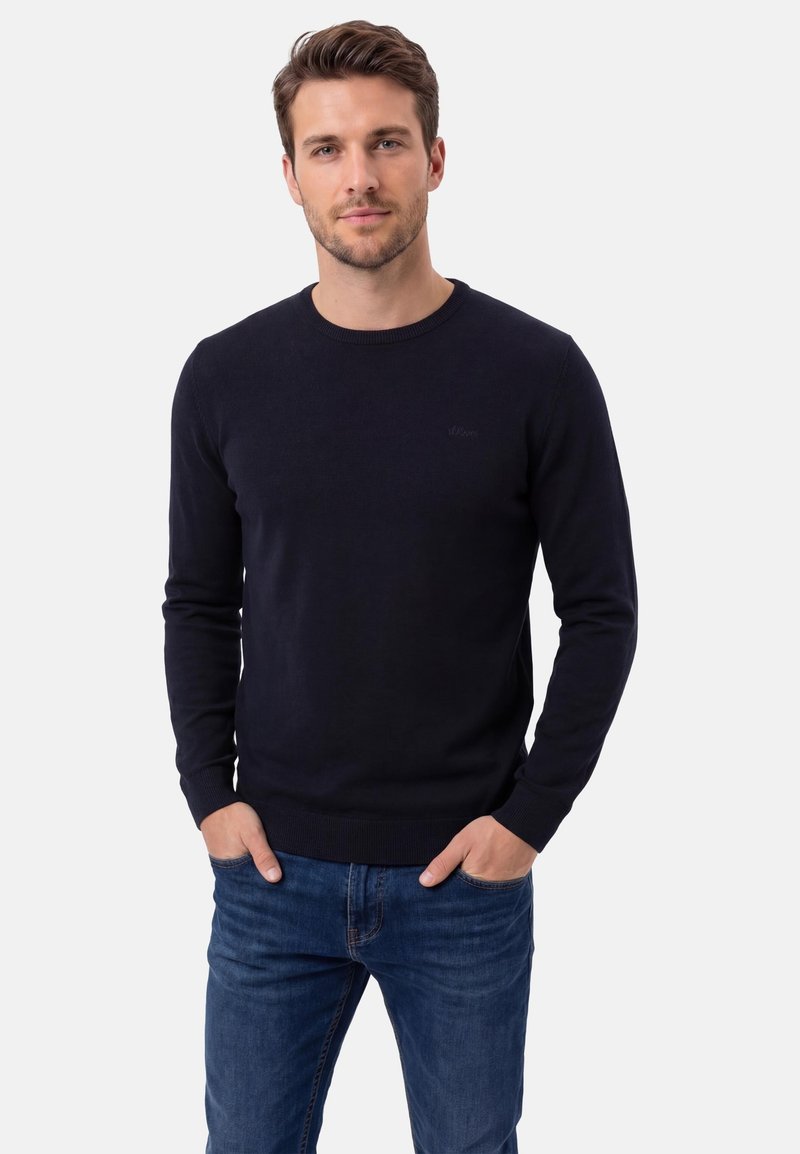 Homme aux cheveux bruns courts portant un pull noir à col rond et un jean bleu, debout les mains dans les poches sur un fond blanc uni.