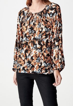 Vrouw draagt een zwarte blouse met lange mouwen met oranje, witte en blauwe bloemen- en vogelprint gecombineerd met een zwarte broek.