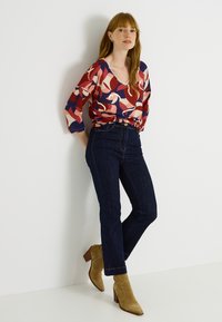Top de manga larga multicolor con patrones abstractos en rojo, rosa y azul marino; combinado con jeans oscuros de cintura alta y botas de tacón beige.