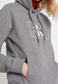 Sudadera con capucha gris con un bolsillo delantero, que presenta un logo blanco dentro de un marco rectangular. Cuerdas con puntas metálicas. Textura de tela suave.