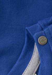 Zbliżenie na niebieską dzianinę z złożonym mankietem zapinanym na metalowy zatrzask z grawerem czterolistnej koniczyny.