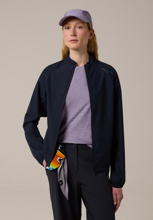 Femme portant une casquette violet clair, une veste zippée noire par-dessus un t-shirt violet clair, un pantalon noir avec des lunettes de soleil accrochées à la ceinture.