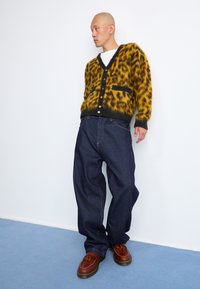 Cardigan i gul og svart leopardmønster, mykt strikket, med knapper foran, kombinert med mørkeblå baggy denimbukser og brune sko med dusk.