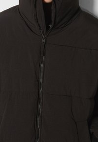 Svart vadderad jacka med hög krage och fullständig framdragkedja; har horisontell sömnad och en slät tygkänsla.