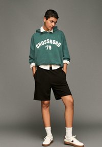 Türkiz oversized kapucnis pulóver fehér "CROSSROAD '79" felirattal, fekete bermuda nadrággal párosítva. Fehér sportcipő barna részletekkel és fehér rövid zoknikkal.