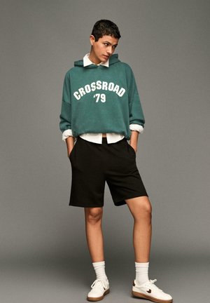 Hoodie oversized color teal con la scritta bianca "CROSSROAD '79", abbinato a pantaloni corti neri. Sneakers bianche con dettagli marroni e calzini bianchi di media lunghezza.
