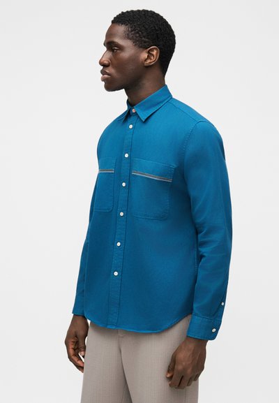 Πουκάμισο σε τιρκουάζ με δύο τσέπες στο στήθος, αντίθετες ραφές και κυρτή βάτα. Το ύφασμα φαίνεται λείο με ελαφριά λάμψη.