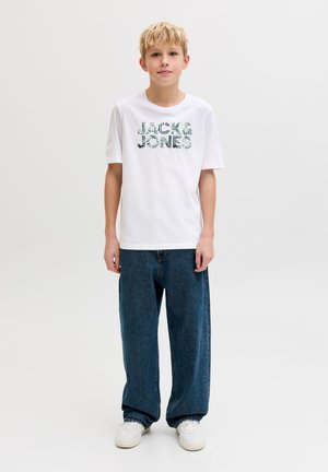 Blond poiss seisab näoga edasi, kandes valget graafilise mustriga t-särki, lahtisi tumelillasid pükse ja valgeid tossusid heleda tausta ees.