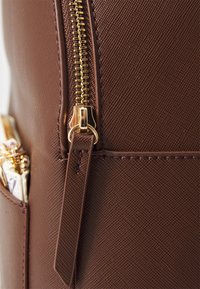 Borsa in tessuto marrone con texture e cerniera dorata, con un primo piano del dettaglio della cerniera e una piccola tasca laterale con accessori in metallo.