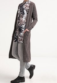 Cardigan lungo con apertura frontale in maglia marrone scuro, indossato sopra una camicia a fantasia floreale, con jeans slim grigi chiari e stivali alla caviglia neri.