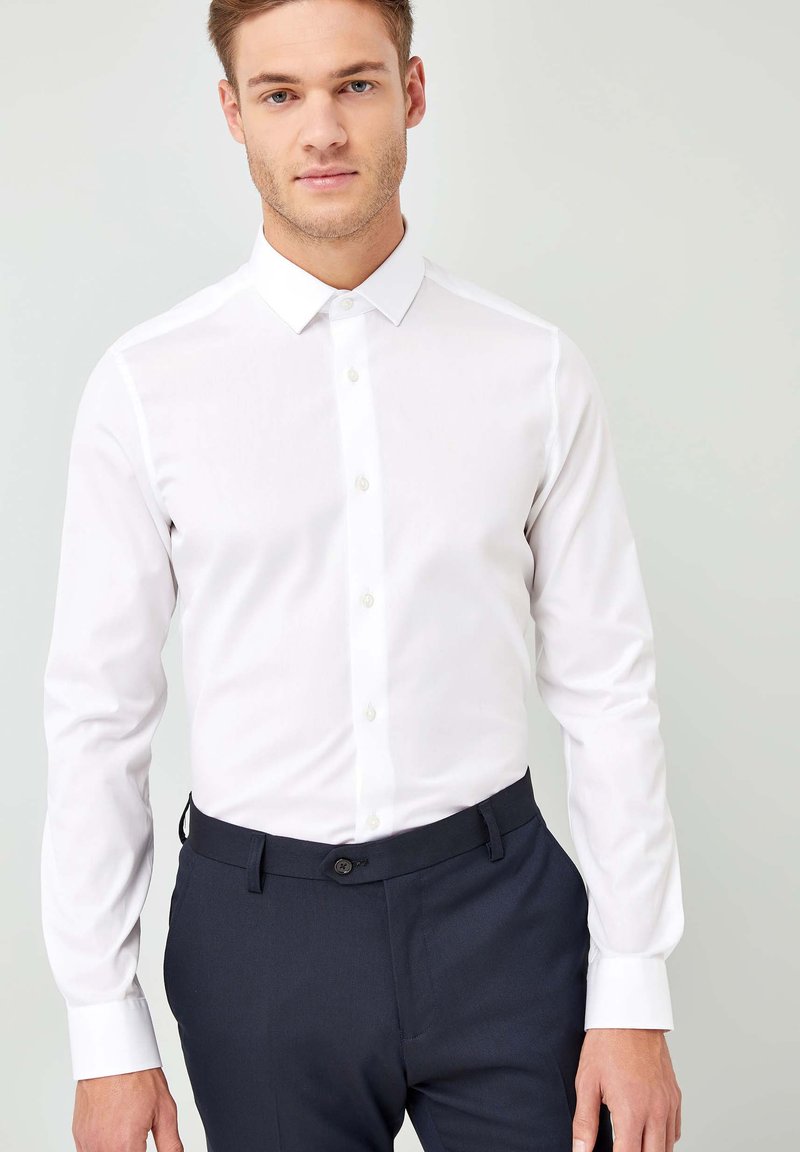Weißes Langarmshirt mit klassischem Kragen, Knopfleiste und glatter Textur, kombiniert mit dunkelblauen Hosen, die eine dezente Textur und einen Knopfverschluss aufweisen.