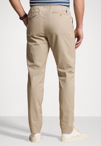 Beiget puuvillahousut rennolla istuvuudella, joustavalla vyötärönauhalla ja kahdella takataskulla. Takataskussa on logo, ja materiaali on sileä.