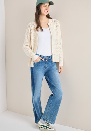 Giovane donna con cappello verde, cardigan crema a maglia aperta, top bianco, jeans blu e sneakers crema con dettagli verdi, in piedi davanti a uno sfondo neutro.