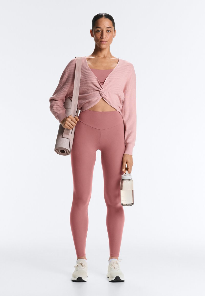 Femme portant un sweat court torsadé rose et un legging, tenant un tapis de yoga roulé sur l'épaule et une bouteille d'eau à la main.