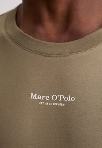 Olijf groene crew neck shirt met witte tekst "Marc O’Polo EST. IN STOCKHOLM" gedrukt op de borst.