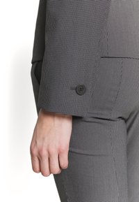 Gros plan d'une personne portant un costume à motif noir et blanc avec un détail de bouton sur la manche et une main visible.