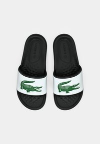Crne natikače s bijelim trakama koje sadrže zeleni krokodil logo, teksturirane umetke za stopala i Lacoste branding unutar.