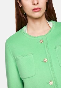 Grüne Filz-Cardigan mit ausgefransten Kanten, zwei vorderen Taschen und dekorativen, blütenförmigen Gold-Ton-Knöpfen. Kurzer, runder Ausschnitt.