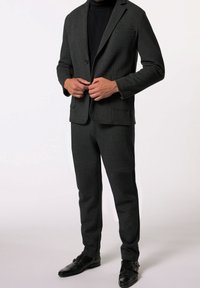 Muž zapíná tmavě šedý blazer na černém roláku s odpovídajícími kalhotami a černými dvojnásobnými botami na přezku na bílém pozadí.