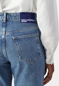 Τζιν παντελόνι σε μεσαίο μπλε χρώμα με ψηλή μέση, διαθέτοντας μια μεγάλη μπλε ετικέτα που γράφει "KARL LAGERFELD JEANS" στην πίσω τσέπη.
