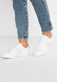 Osoba nosząca niebieskie dżinsy i białe trampki Lacoste z różowozłotymi akcentami oraz powtarzającym się tekstem marki po bokach, stojąca na białej podłodze.