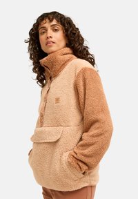 Femme aux cheveux bouclés portant une veste polaire sherpa beige et brun clair à col montant et poche avant, se tenant devant un fond uni.