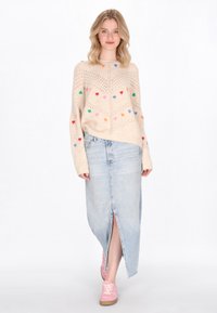 Maglione lavorato a maglia beige con motivi a cuore multicolori, abbinato a una gonnella di jeans chiari con uno spacco anteriore e sneakers rosa.