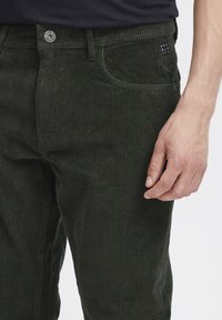 Donkergroene corduroy broek met verticale ribbels. Voorzien van een knoopsluiting, een voorzak en een merkpatch op de tailleband.
