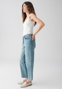 Witte tanktop met dunne bandjes, lichtblauwe hoog gewaagde jeans en beige instap schoenen. Model poseert in zijprofiel tegen een neutrale achtergrond.