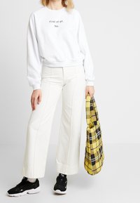 Sweat-shirt blanc avec du texte noir, large pantalon crème, et baskets noires. Porte un sac à carreaux jaune avec des accents noirs et gris.