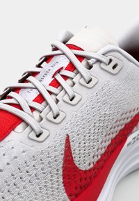 Chaussure de course Nike Pegasus avec une tige en mesh blanche ornée d'accents rouges, des lacets gris et une semelle intermédiaire lisse. Logo swoosh remarquable en rouge.