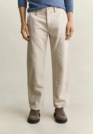 Personne portant un pantalon droit beige et des bottes marron à lacets, debout devant un fond uni clair.