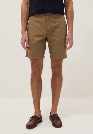 Homme portant un short beige jusqu’aux genoux, ceinture noire, chemise noire et chaussures bateau marron foncé à lacets, debout sur un fond uni.