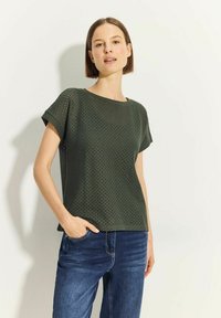 Mørkegrøn kortærmet top med et tekstureret, perforeret design. Rund halsudskæring og afslappet pasform, parret med blå denimjeans.