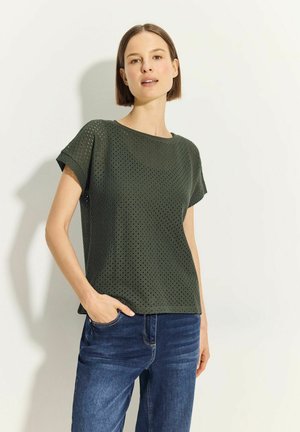 Tmavě zelený top s krátkým rukávem a texturovaným, perforovaným designem. Kulatý výstřih a uvolněný střih, doplněný modrými džínami.