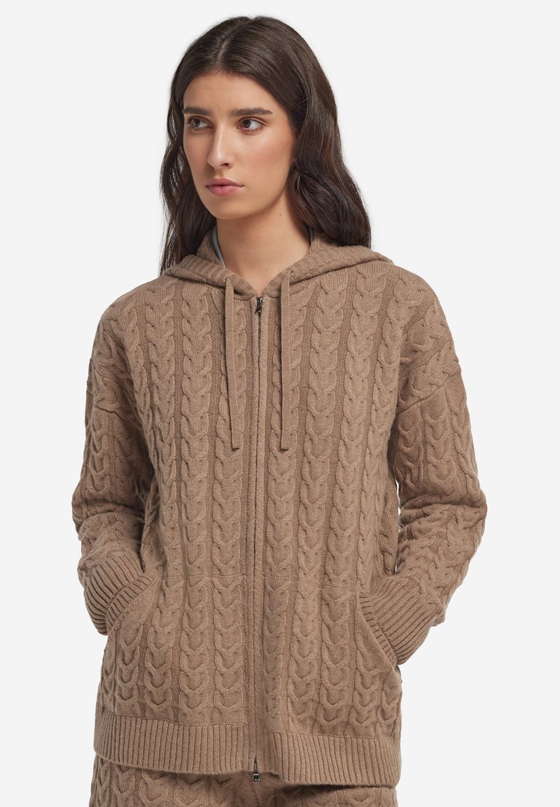Femme portant un sweat à capuche beige en maille torsadée avec fermeture éclair et poches, debout les mains dans les poches, regardant sur le côté sur un fond blanc.