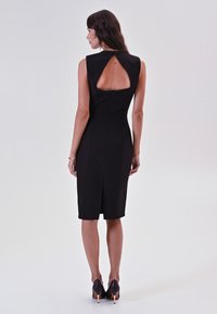 Frau, die mit dem Rücken zur Kamera steht und ein enges, schwarzes ärmelloses Kleid mit einem dreieckigen Rückenausschnitt sowie schwarzen High Heels trägt.