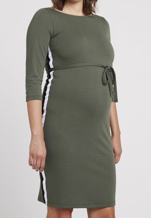 Kobieta w ciąży w dopasowanej oliwkowej sukience z rękawami do łokcia i białym paskiem po bokach, wiązana w talii.