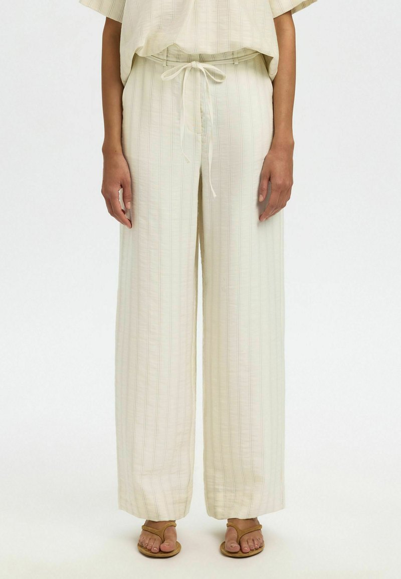 Beige bukser med vide ben og subtile lodrette striber, bundet i taljen, båret med brune sandaler med stropper og matchende løs top.