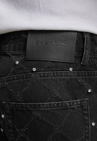 Tuvumā redzami melni džinsi ar teksturētu dimanta rakstu, maziem rhinestone rotājumiem un melnu ādas plāksni ar iepludinātu zīmolu.