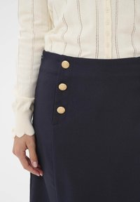 Jūras spēku svārki ar gludu tekstūru, ar četriem zelta pogām sānos un diskrētu kabatas detaļu. Stilizēti ar krēmveida adītu topu.