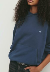 Persona che indossa un felpa blu navy con un piccolo logo bianco, una cintura nera borchiata e pantaloni neri, in posa con una mano in tasca.
