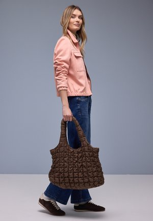 Donna con giacca rosa, jeans blu e sneakers che cammina portando una grande borsa trapuntata marrone su uno sfondo semplice.