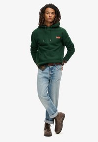 Felpa verde con cappuccio, tasca frontale e cordini, abbinata a jeans leggeri blu strappati e stivali marroni con lacci.