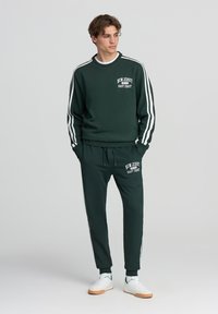 Completo felpa e pantaloni verde scuro con strisce bianche, caratterizzato da testo stampato "NEW JERSEY EAST COAST." Il materiale sembra morbido e con texture.