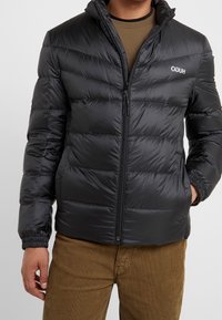 Mann trägt eine schwarze gesteppte Jacke mit Reißverschluss und Händen in den seitlichen Taschen, kombiniert mit braunen Cordhosen.