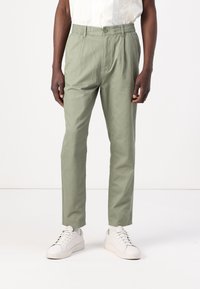 Mężczyzna w oliwkowozielonych casualowych spodniach i białych sneakersach, stojący na tle jednolitego białego tła.