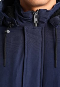 Nahaufnahme einer Person, die eine dunkelblaue Jacke mit Reißverschluss trägt, schwarz gefütterte Kordeln und ein Fleecefutter am Kragen sichtbar.