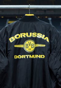 Veste bomber noire avec texte jaune "Borussia Dortmund" et logo BVB 09 suspendue à une barre métallique.